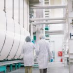 Back view full length portrait of two factory workers wearing lab coats crossing production workshop, copy space